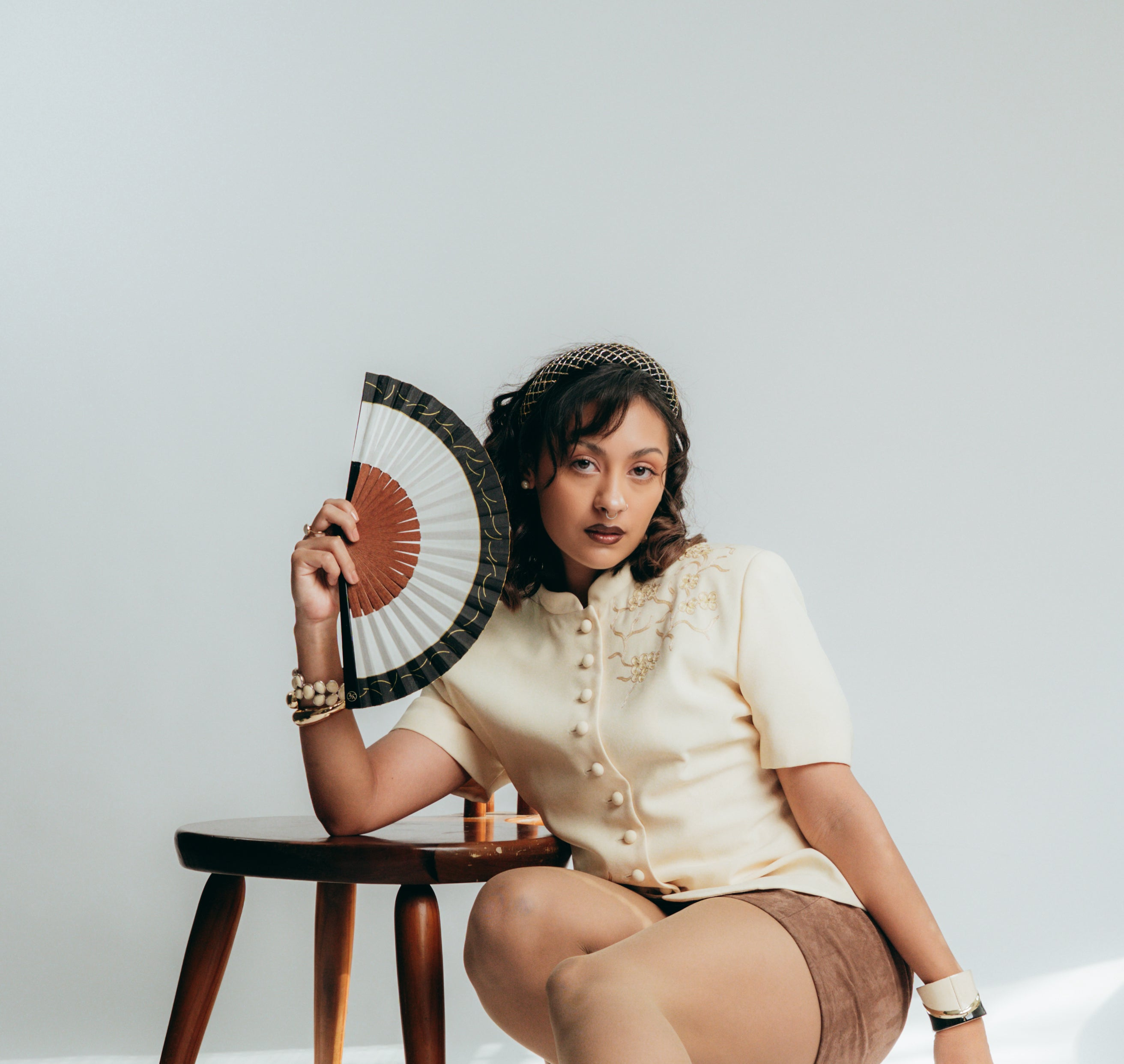 Woman holding a fan, sitting on a chair against a plain background